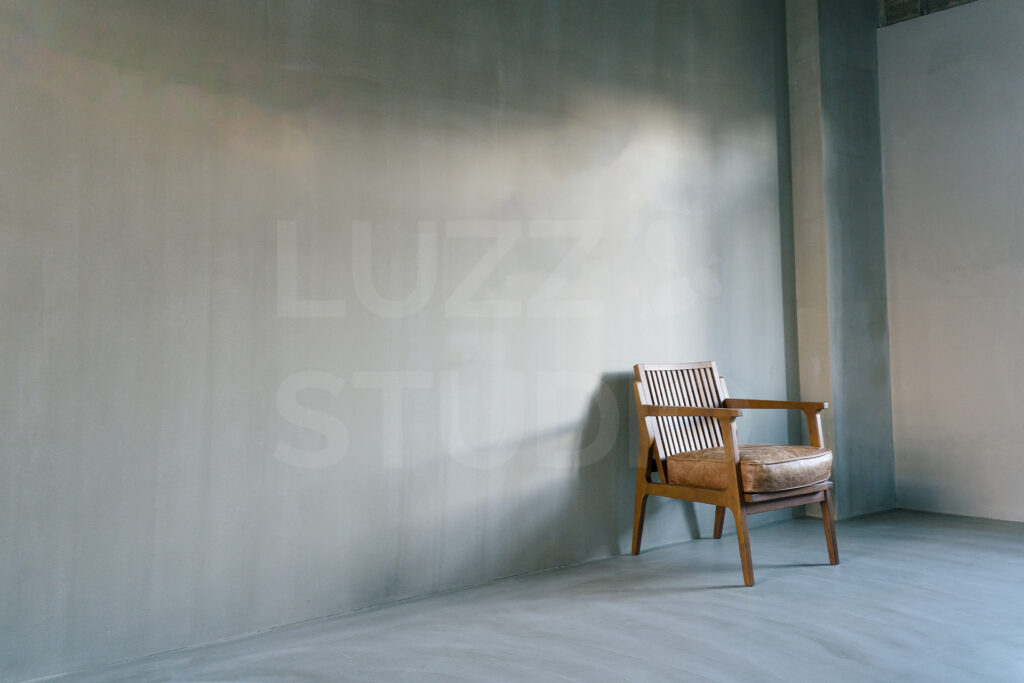 大阪の撮影会社によるスタジオ撮影サービス。質感のあるグレーのモルタル壁を背景に、ヴィンテージチェアと柔らかな自然光を活かした、洗練されたポートレート・広告撮影