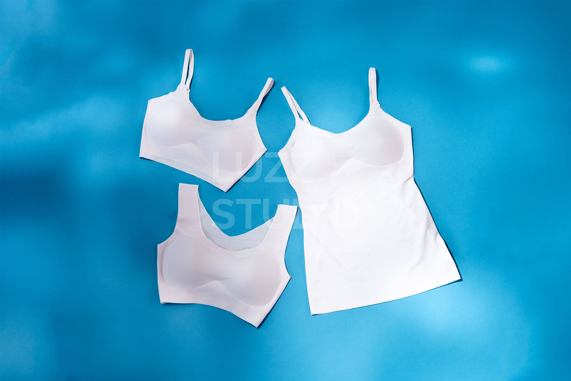 青色の背景紙の上にライトピンクのブラジャー、キャミソール、スポーツブラを配置した商品撮影写真。光の陰影による春夏のさわやかな印象をプラスしている