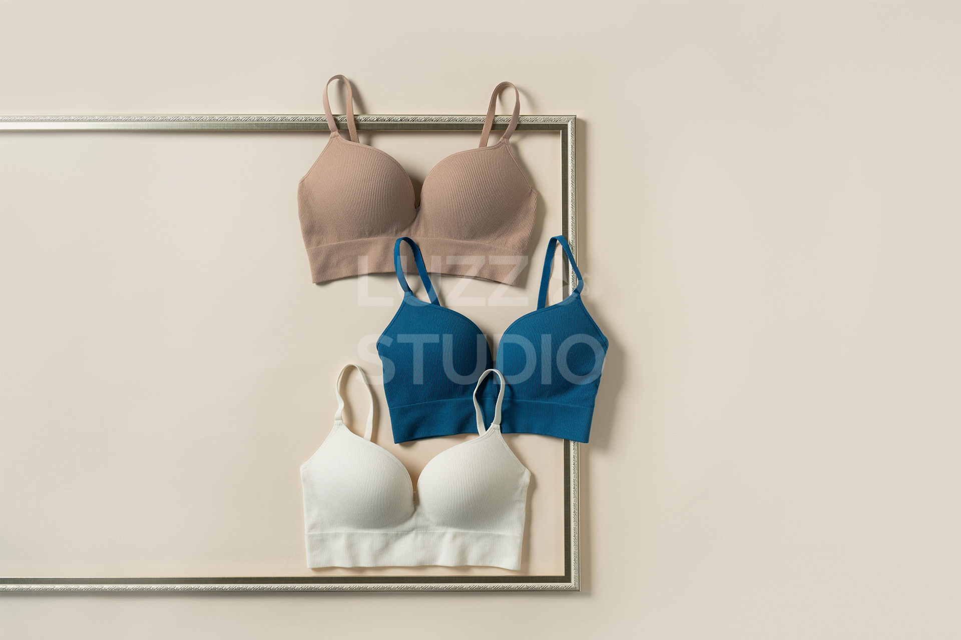 ベージュの背景紙の上にベージュ、ブルー、ホワイトのブラジャーを配置した商品撮影写真。ゴールドの額縁の上に配置することで、女性らしさを演出している。