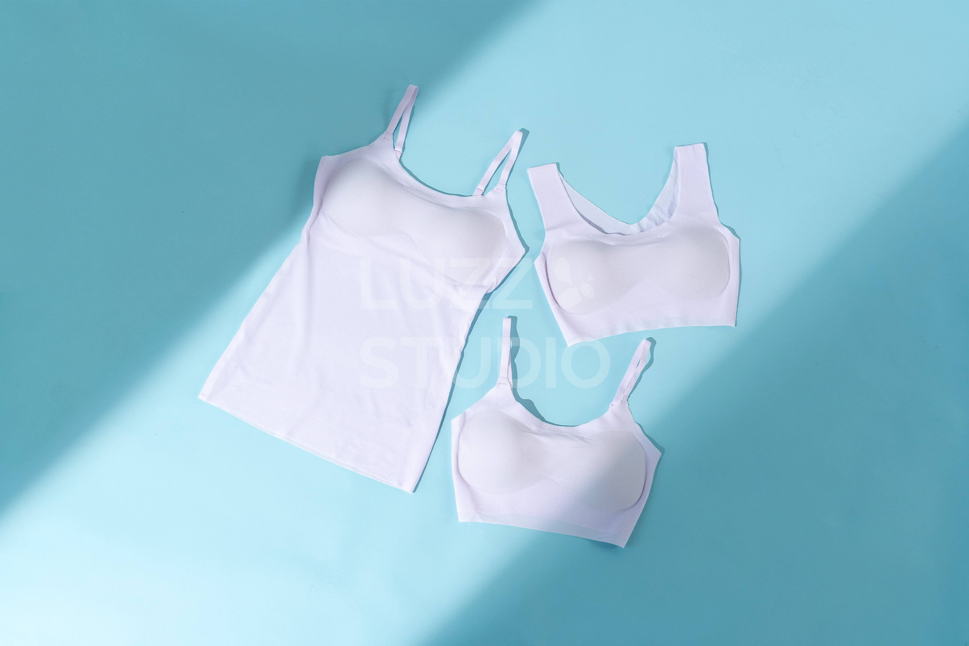 青色の背景紙の上にピンクのブラジャー、キャミソール、スポーツブラを配置した商品撮影写真。光の筋による陰影で商品にフォーカスされる工夫がされている。