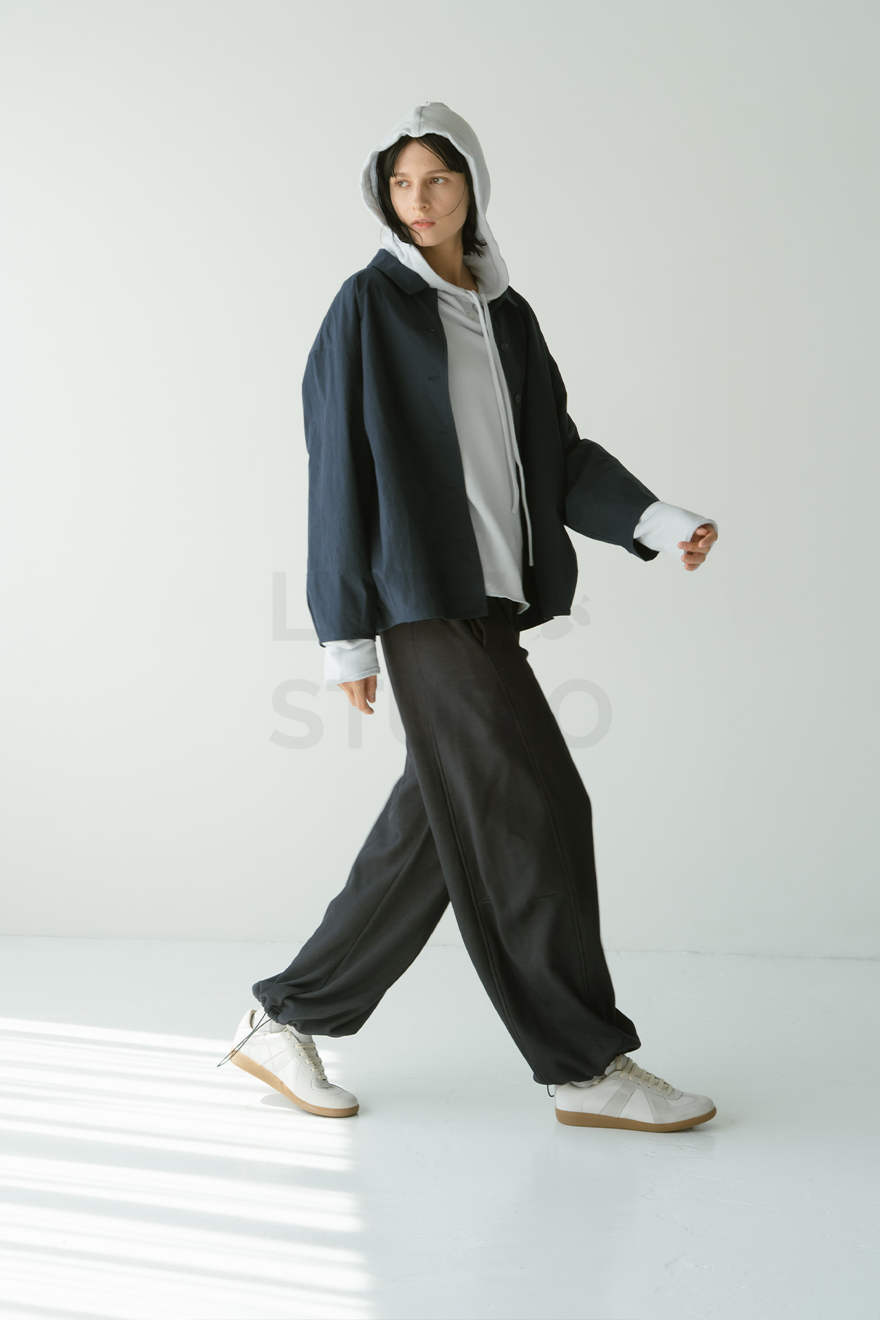フード付きトップスにダークネイビーのジャケットとワイドパンツを合わせ、白いスニーカーを履いてスタジオ内を軽やかに歩くモデルの全身ショット